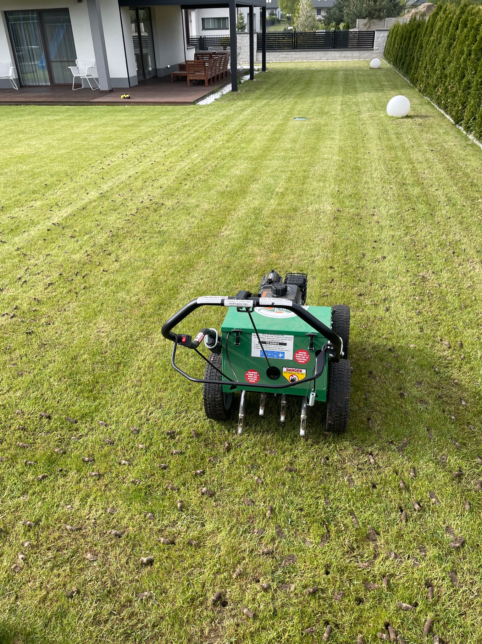 Zielony aerator do trawy na trawniku, widoczne rdzenie wyciągnięte z gleby, w tle nowoczesny dom z tarasem i zadbany ogród.