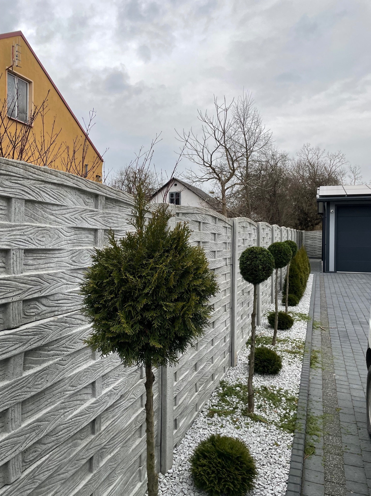 Ogród z ozdobnymi, formowanymi krzewami wzdłuż betonowego, ażurowego ogrodzenia, wysypany białym grysem, widok na garaż i dom w tle.