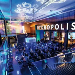 Widok z góry na salę konferencyjną Hydropolis z publicznością siedzącą na krzesłach, prelegentów na scenie i dekoracyjnym oświetleniem sufitowym.