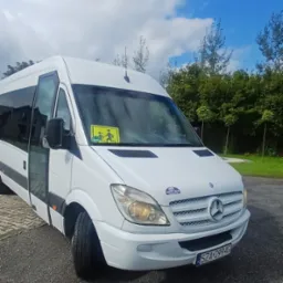 Biały bus Mercedes-Benz z otwartymi drzwiami, naklejką 'dzieci' na szybie i logo firmy 'MATTI-BUS' na masce, zaparkowany na żwirowym parkingu w słoneczny dzień.