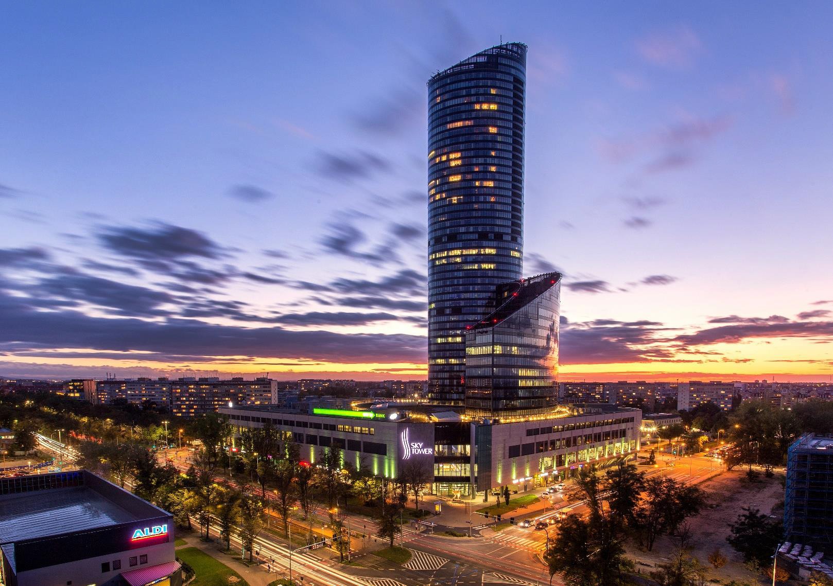 Wieczorny widok na Sky Tower we Wrocławiu z długą ekspozycją świateł ulicznych i zachodem słońca w tle.