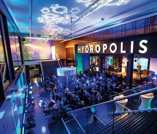 Widok z góry na salę konferencyjną Hydropolis z publicznością siedzącą na krzesłach, prelegentów na scenie i dekoracyjnym oświetleniem sufitowym.