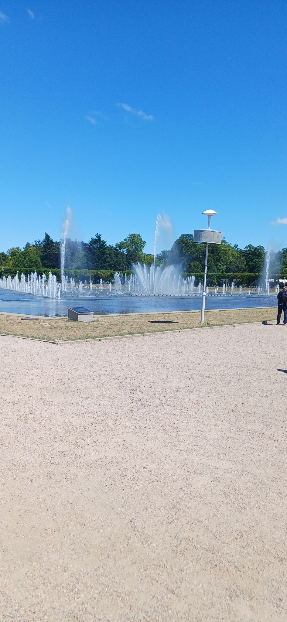 Widok na fontannę w parku, w tle drzewa i błękitne niebo, na pierwszym planie żwirowa ścieżka.