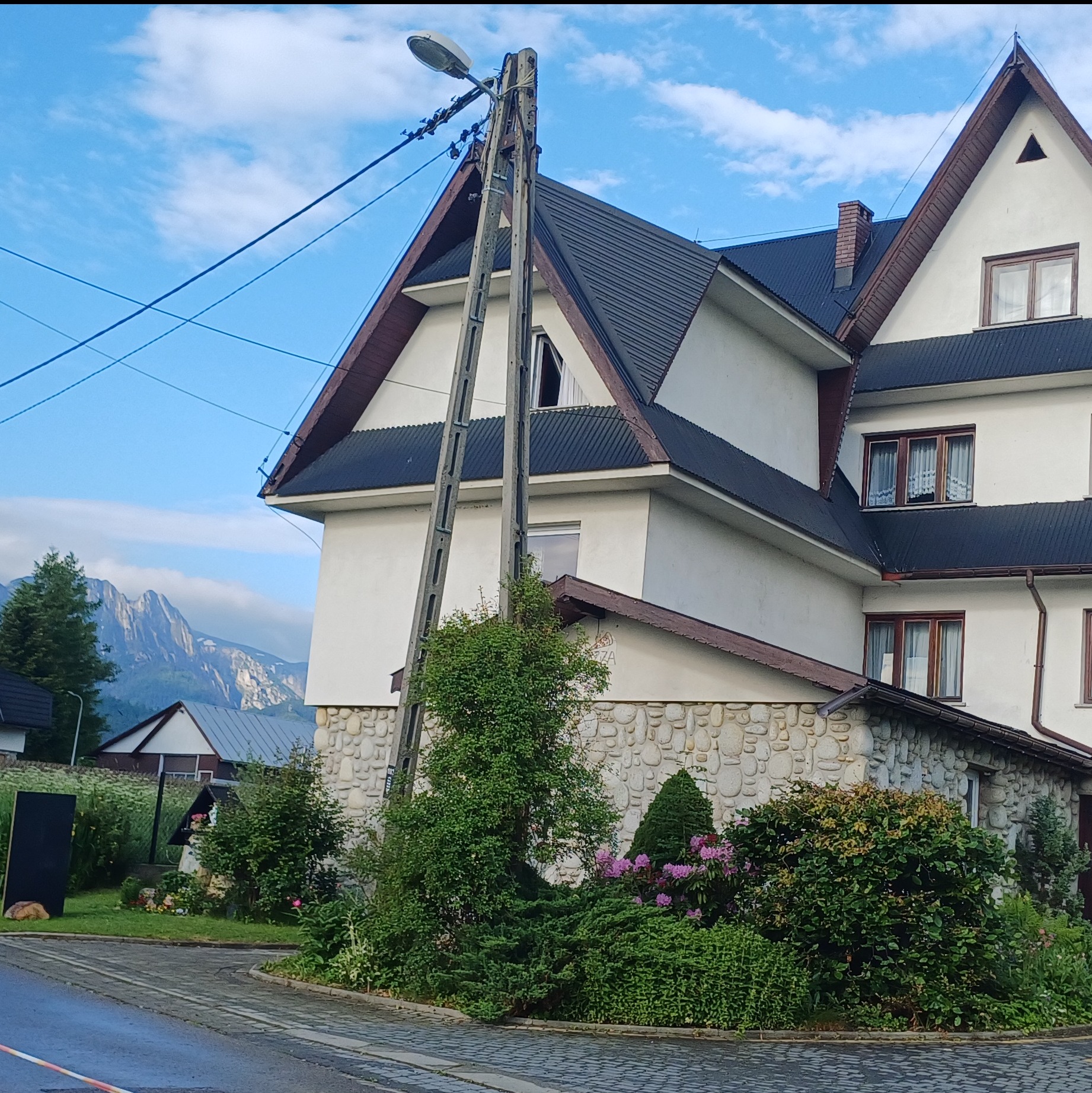 Dwupiętrowy budynek z kamienną elewacją na parterze, ciemnym dachem i drewnianymi obramowaniami okien, z widokiem na góry w tle i słupem elektrycznym na pierwszym planie.