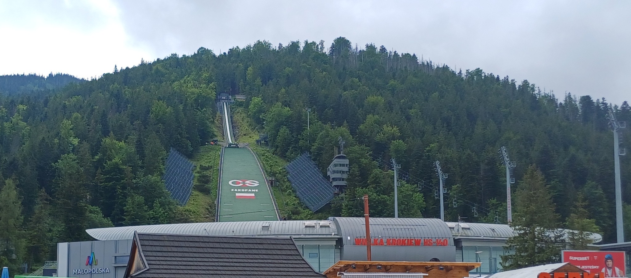 Panorama Wielkiej Krokwi w Zakopanem, widok na skocznię narciarską z zielonym igelitem, las porastający zbocze góry i budynki w tle, pochmurne niebo.