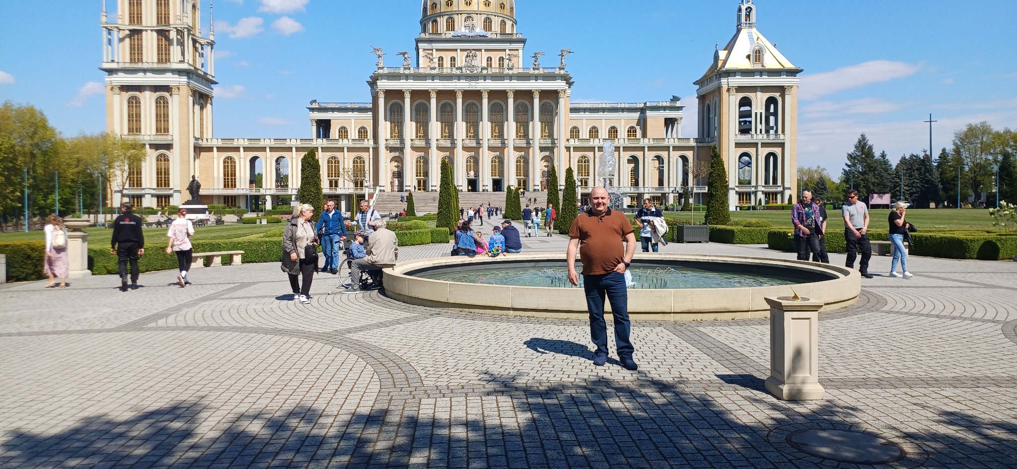 Sanktuarium w Licheniu Starym w słoneczny dzień, mężczyzna w brązowej koszulce polo i jeansach stoi na pierwszym planie przed fontanną, w tle widoczni spacerujący ludzie.