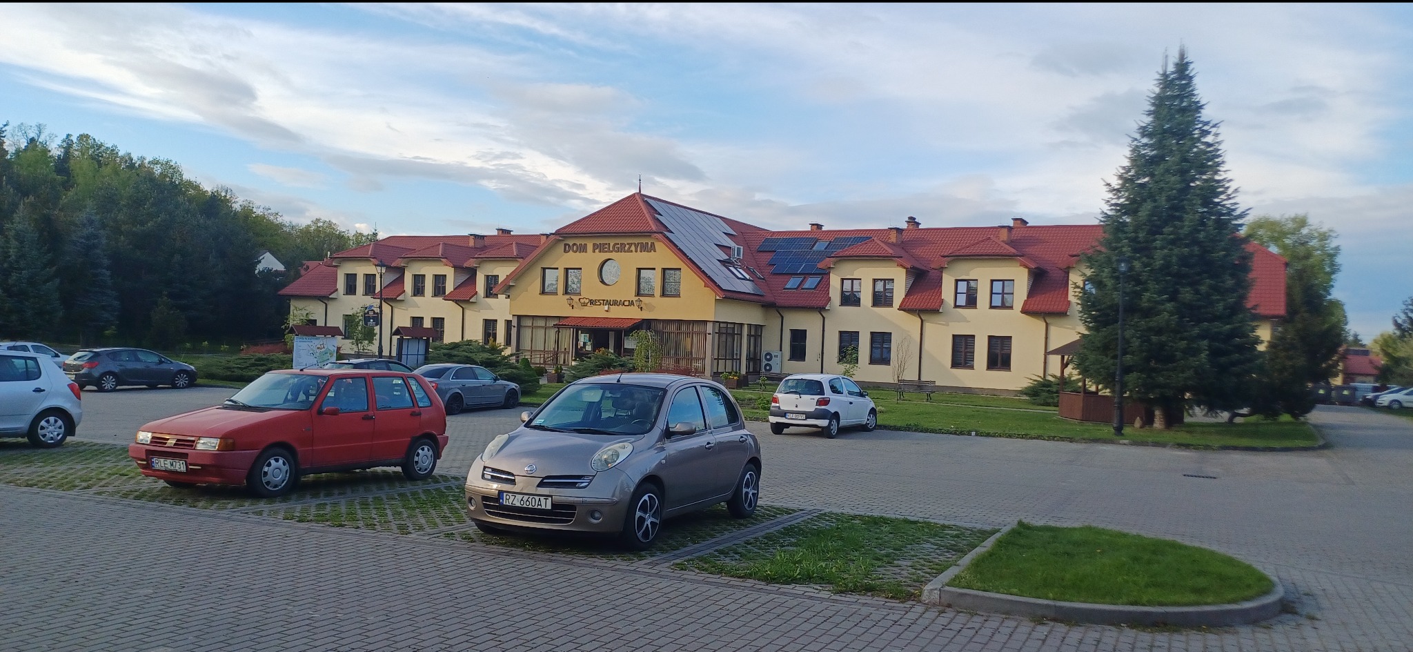 Parking przed budynkiem Domu Pielgrzyma z restauracją, widoczne zaparkowane samochody osobowe i panele słoneczne na dachu.