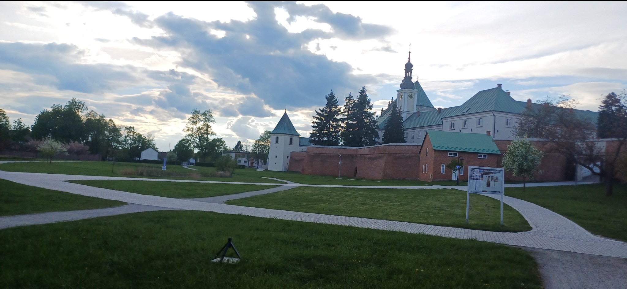 Widok na klasztor z zielonymi dachami i wieżą zegarową, otoczony murem z czerwonej cegły, z ścieżkami spacerowymi i trawnikami na pierwszym planie, pochmurne niebo w tle.