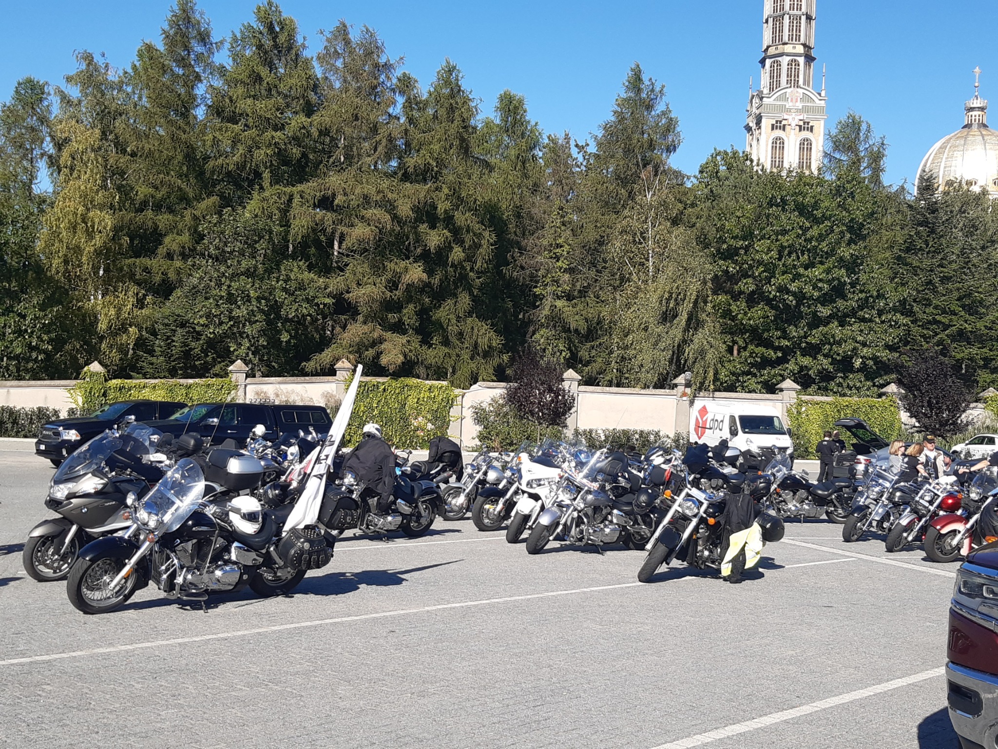 Grupa motocykli zaparkowana na placu, w tle drzewa i wieża kościoła w Licheniu Starym, Polska, słoneczny dzień.