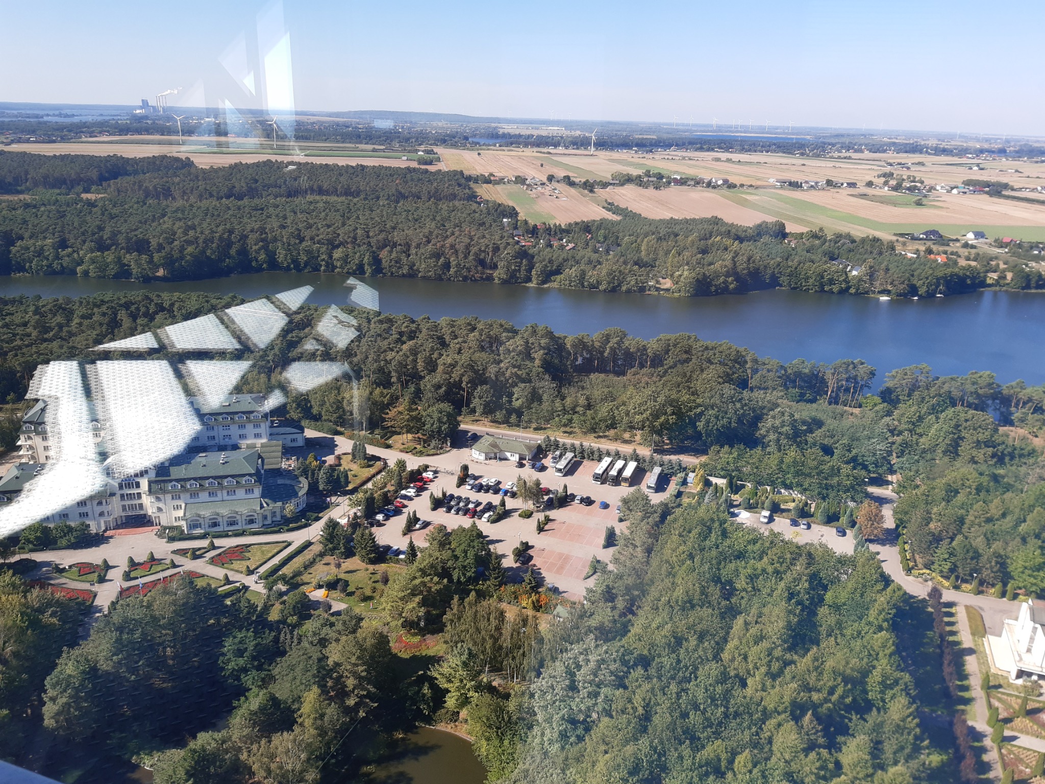 Widok z góry na parking z zaparkowanymi busami i samochodami osobowymi przy jeziorze, obok hotelu z zadbanym ogrodem, w tle pola uprawne i elektrownia w oddali.