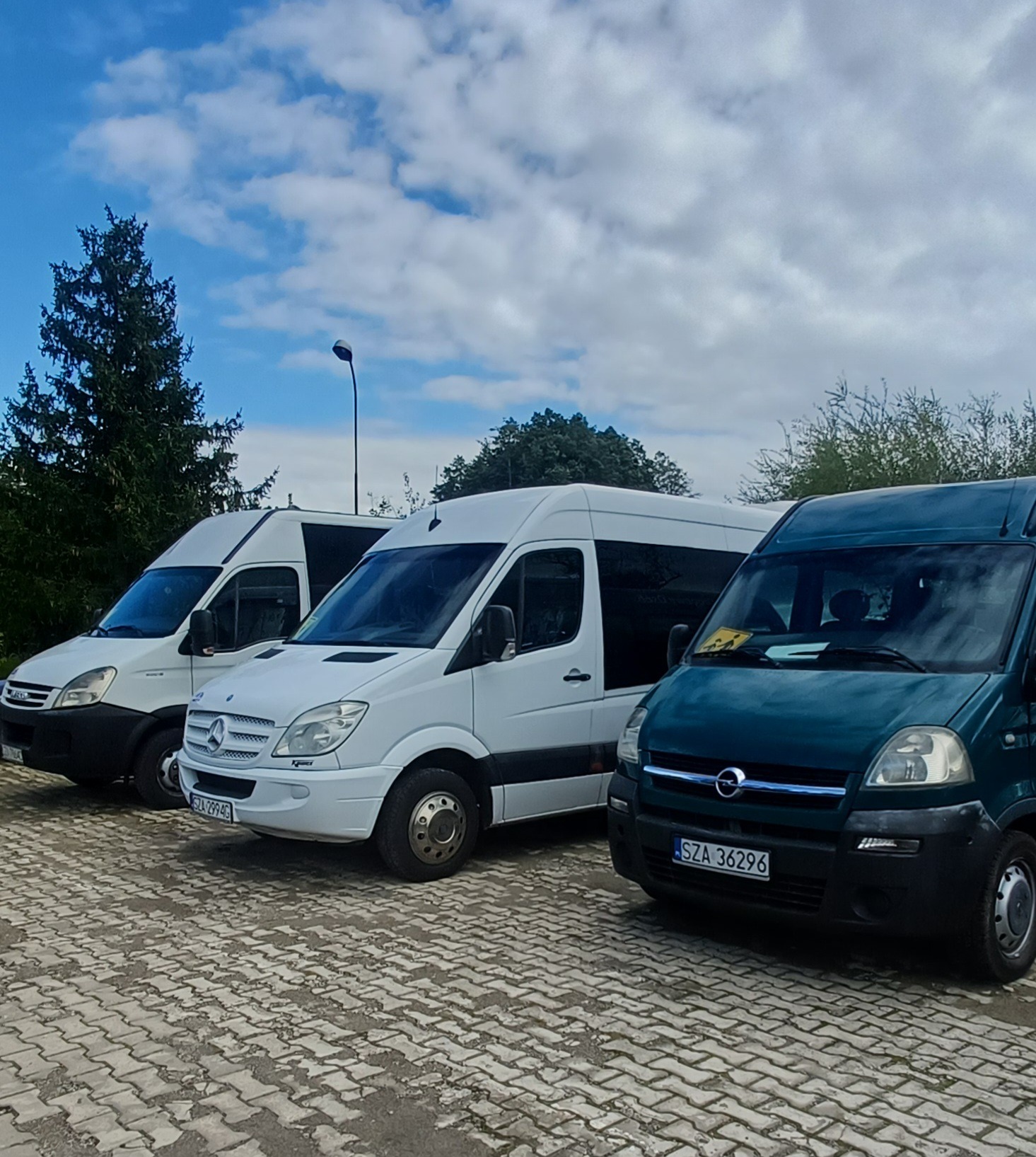 Trzy busy: biały Iveco, biały Mercedes Sprinter i ciemnozielony Opel Movano, zaparkowane na brukowanym placu, z drzewem i pochmurnym niebem w tle.