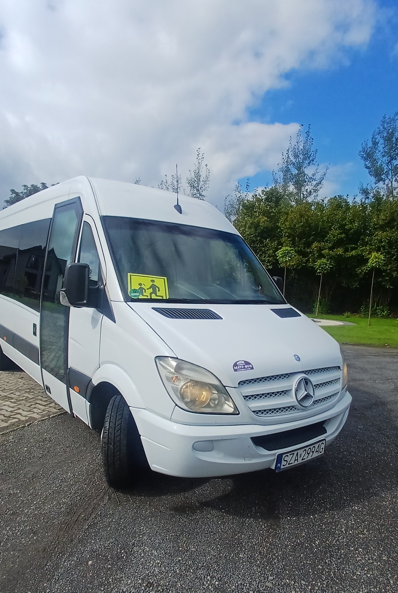 Biały bus Mercedes-Benz z otwartymi drzwiami, naklejką 'dzieci' na szybie i logo firmy 'MATTI-BUS' na masce, zaparkowany na żwirowym parkingu w słoneczny dzień.