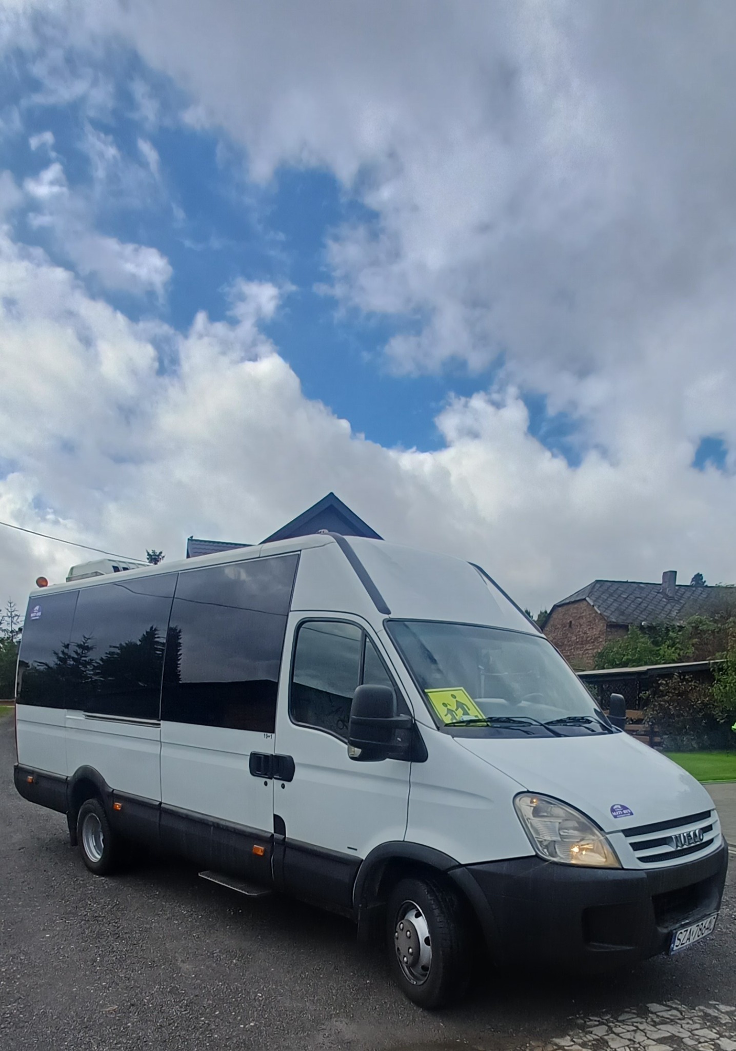 Biały bus Iveco z przyciemnianymi szybami, oznaczony żółtą tabliczką 'dzieci', zaparkowany na tle pochmurnego nieba i zabudowań.