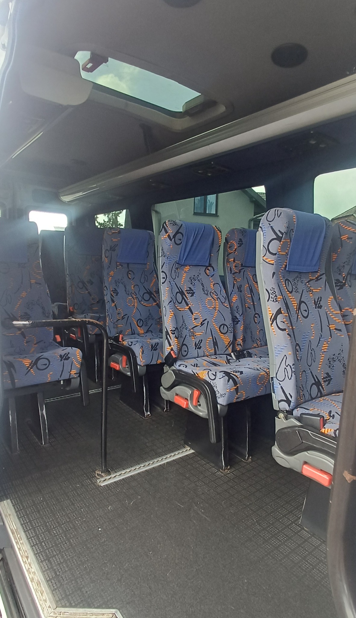 Wnętrze busa z rzędami foteli w niebieskiej tapicerce ze wzorem, widok na okno dachowe i fragment zabudowań na zewnątrz.