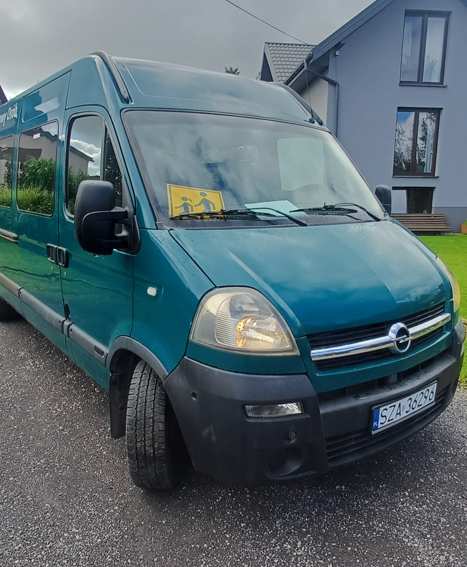 Zielony bus w Zawierciu z żółtą naklejką 'dzieci' na szybie, zaparkowany przed domem. Widok na przód pojazdu i otoczenie.