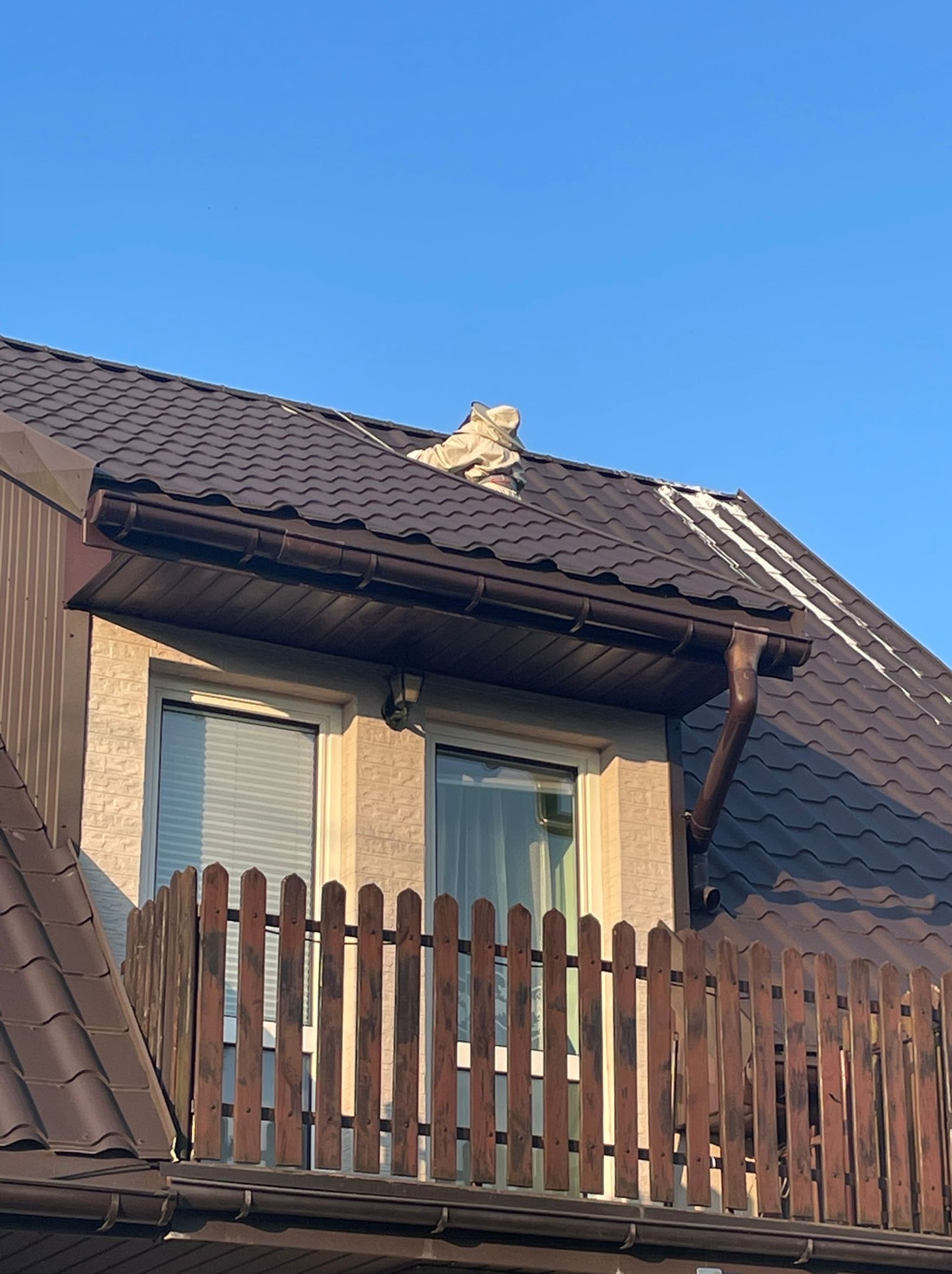 Prace na dachu domu z brązową dachówką, widoczne zabezpieczenia i przygotowania do naprawy lub malowania, brązowa rynna i drewniana balustrada balkonu.