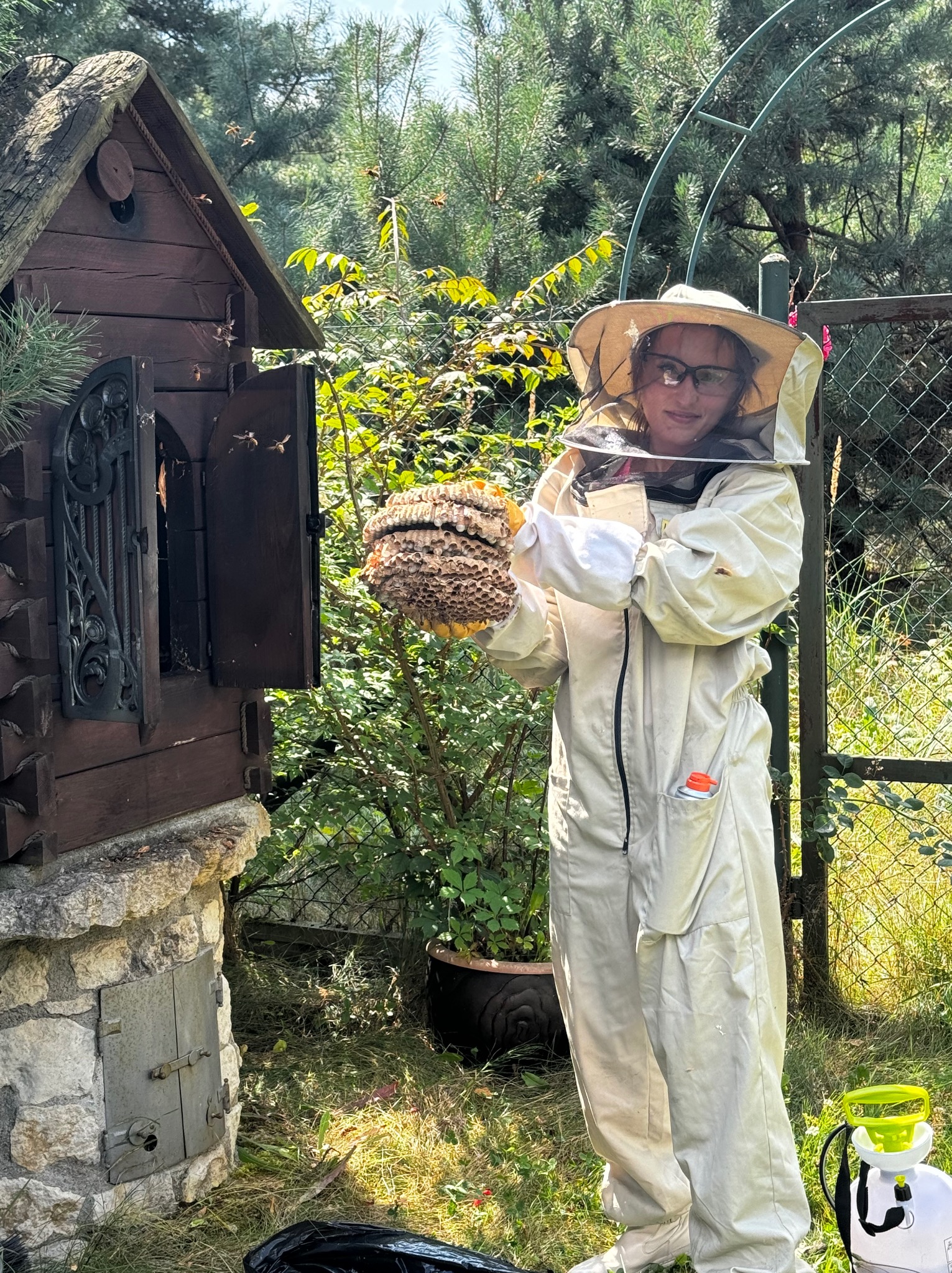 Osoba w kombinezonie ochronnym i siatce na twarzy trzyma w dłoniach duży plaster pszczeli przed drewnianym domkiem dla owadów, z którego wylatują pszczoły, w tle zielona roślinność i ogrodzenie...