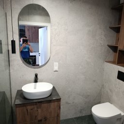 Ivan Kurtiak - Nowoczesna łazienka z owalnym lustrem odbijającym fragment kuchni, umywalką nablatową i drewnianymi półkami. Szare płytki imitujące kamień.