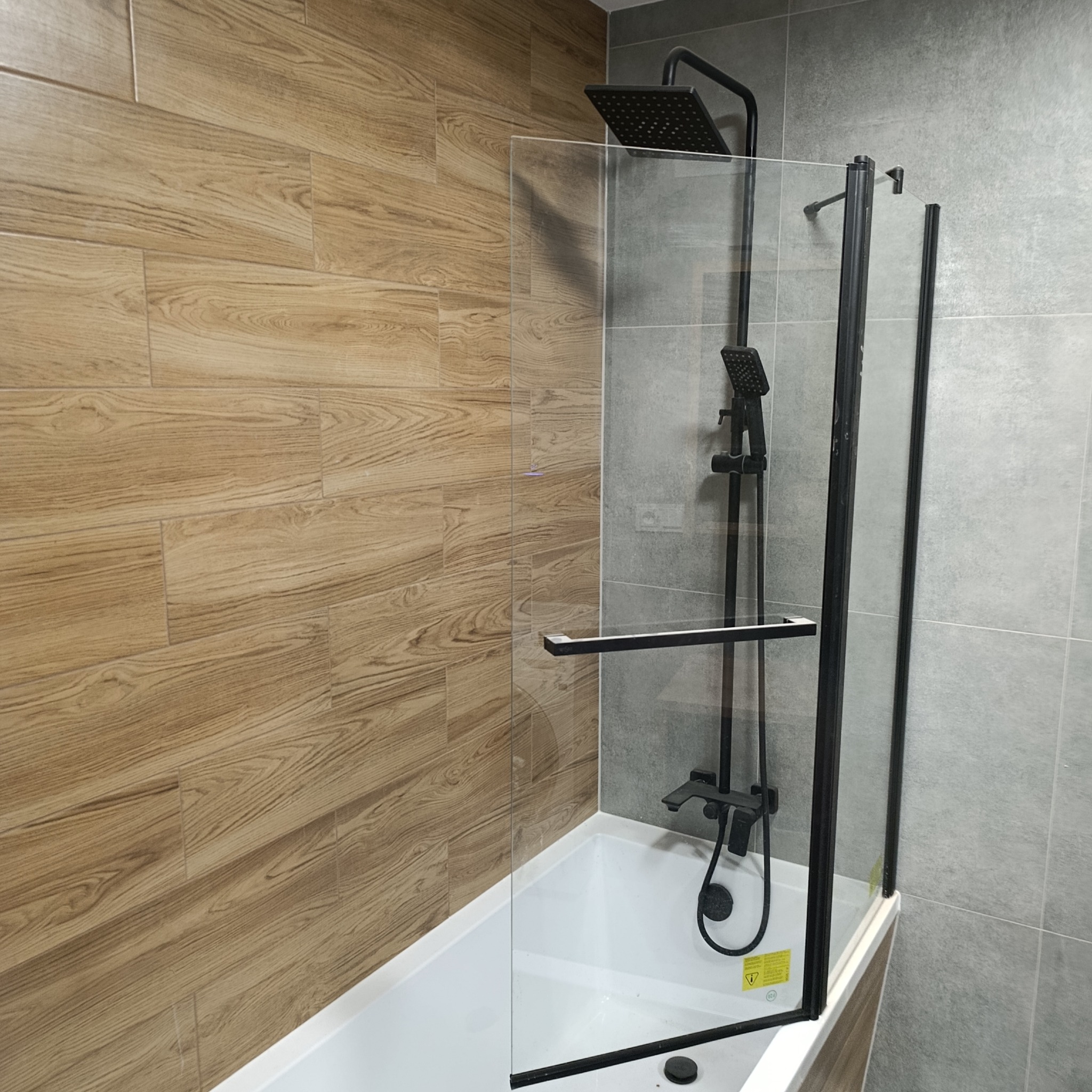 Nowoczesna łazienka po remoncie: biała wanna z czarną armaturą i szklanym parawanem, drewnopodobne i szare płytki na ścianach.