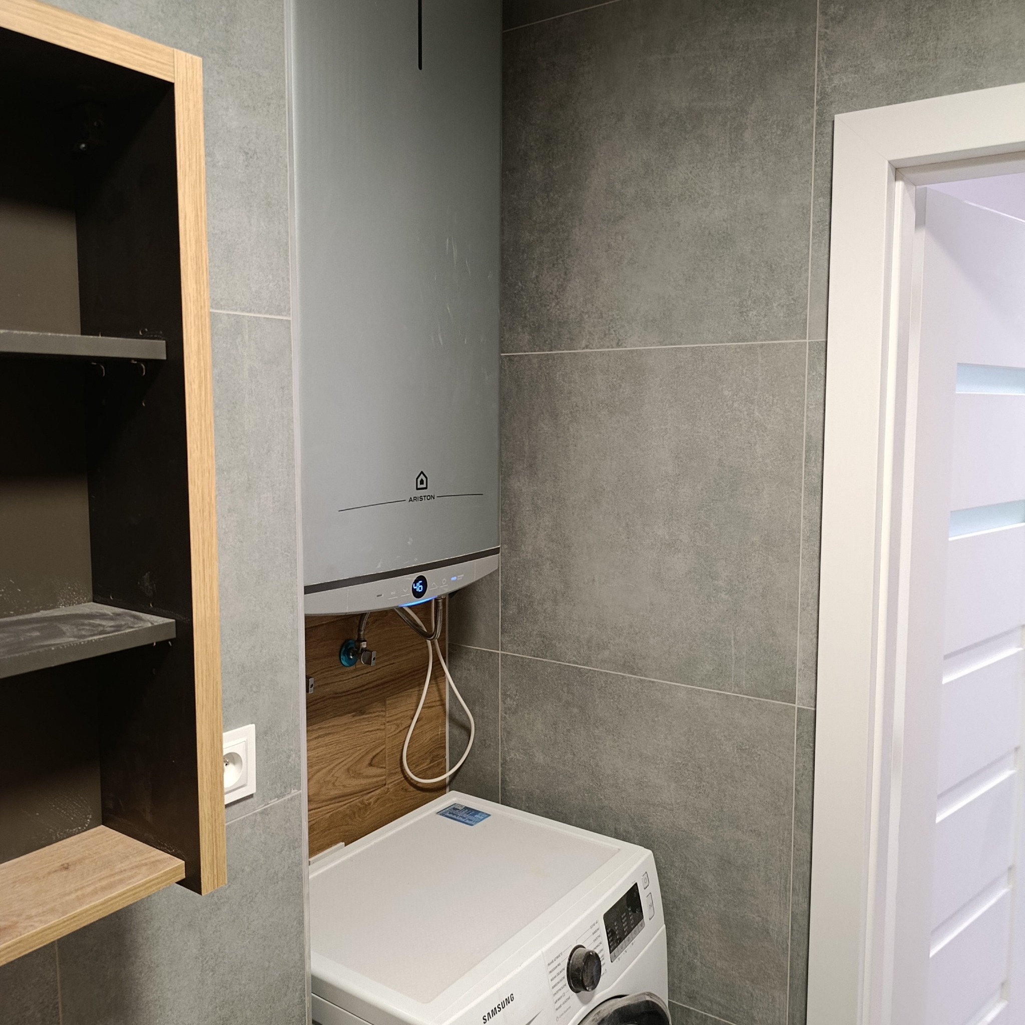 Nowoczesna łazienka z szarymi płytkami, pralką Samsung i wiszącym bojlerem Ariston. Widoczny fragment szafki z drewnianymi półkami i białe drzwi z przeszkleniem.
