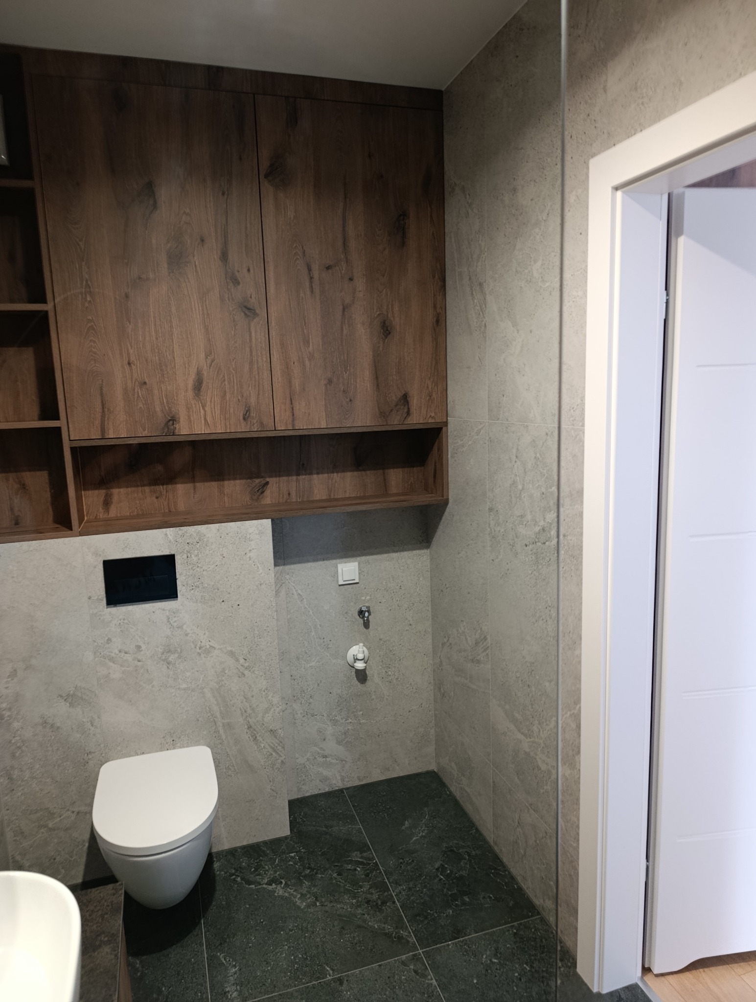 Nowoczesna łazienka po remoncie: szare płytki imitujące kamień, podwieszana toaleta i drewniane szafki wiszące. Minimalistyczny design, widoczne detale wykończenia.