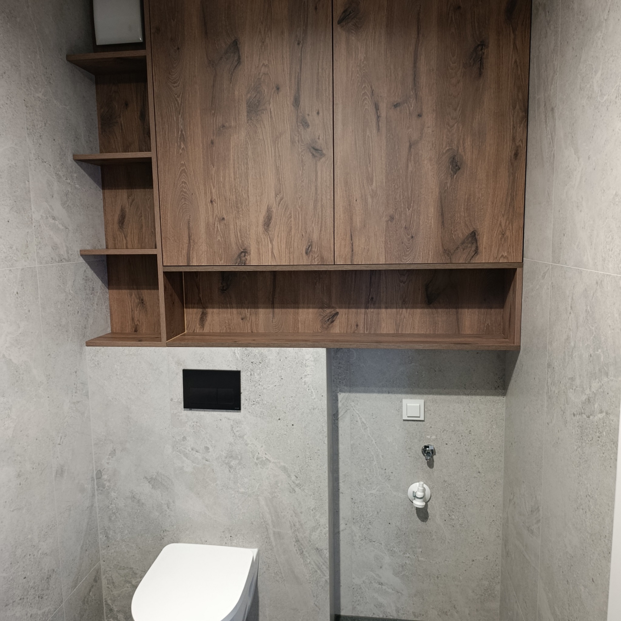 Nowoczesna łazienka z szarą płytką imitującą kamień, drewnianymi szafkami wiszącymi i podtynkową toaletą. Minimalistyczny design, oświetlenie punktowe.