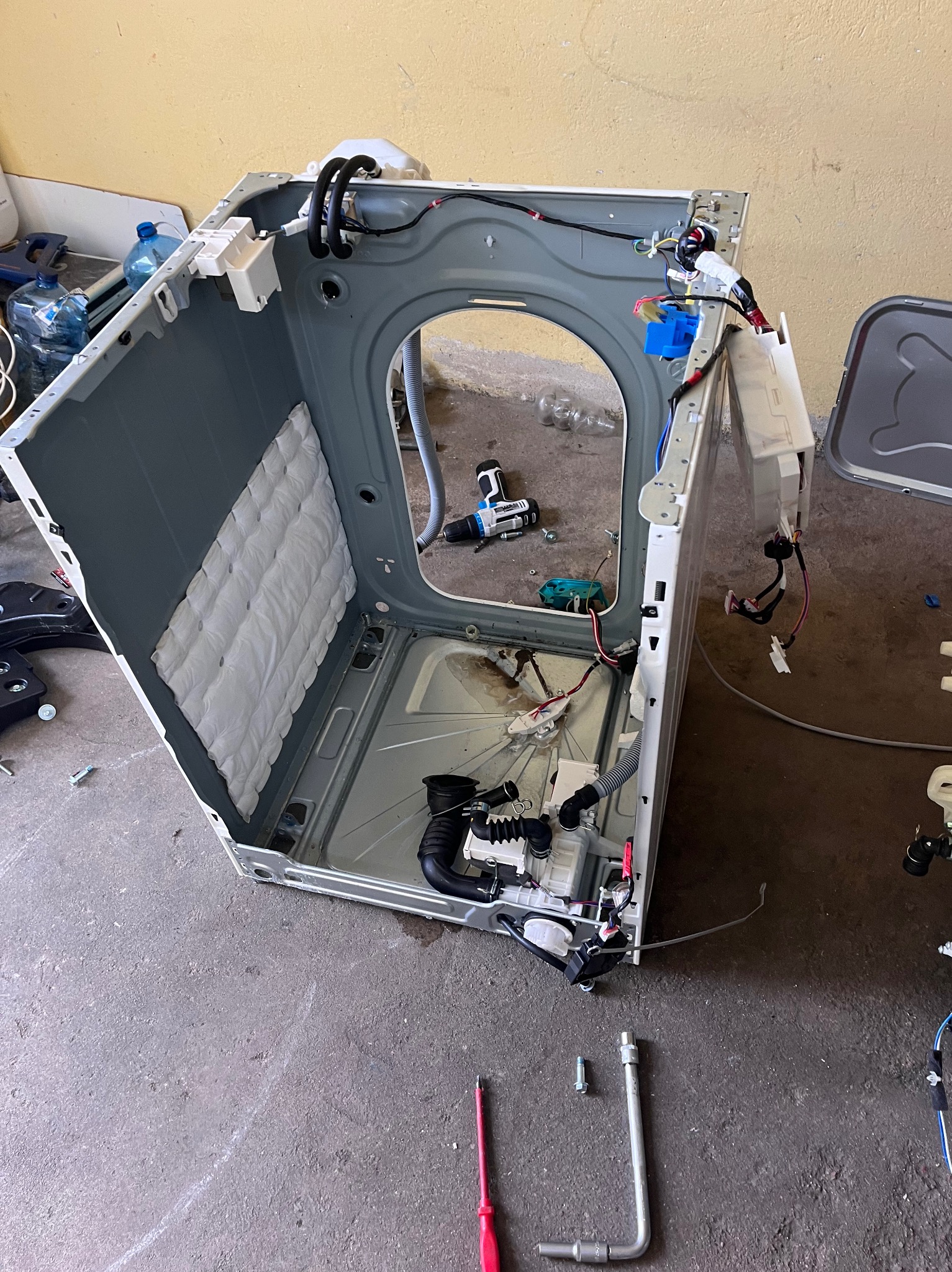 Rozłożona pralka automatyczna podczas naprawy, widoczne wnętrze obudowy, elementy elektroniczne, narzędzia: wkrętarka akumulatorowa, śrubokręt i klucz.