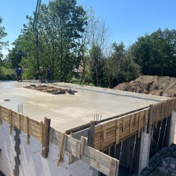 KHRYSTYNA KALYNIAK KALYNIAK BUD - Wylewanie betonu na strop domu z białych bloczków, widoczna pompa do betonu i pracownicy w tle, szalunek z desek na krawędzi stropu.