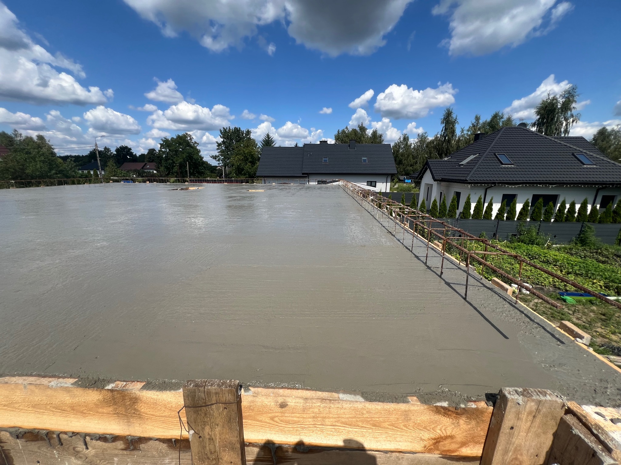 Świeżo wylana, gładka płyta fundamentowa z widocznym zbrojeniem obwodowym, na tle zabudowy jednorodzinnej i błękitnego nieba z chmurami.