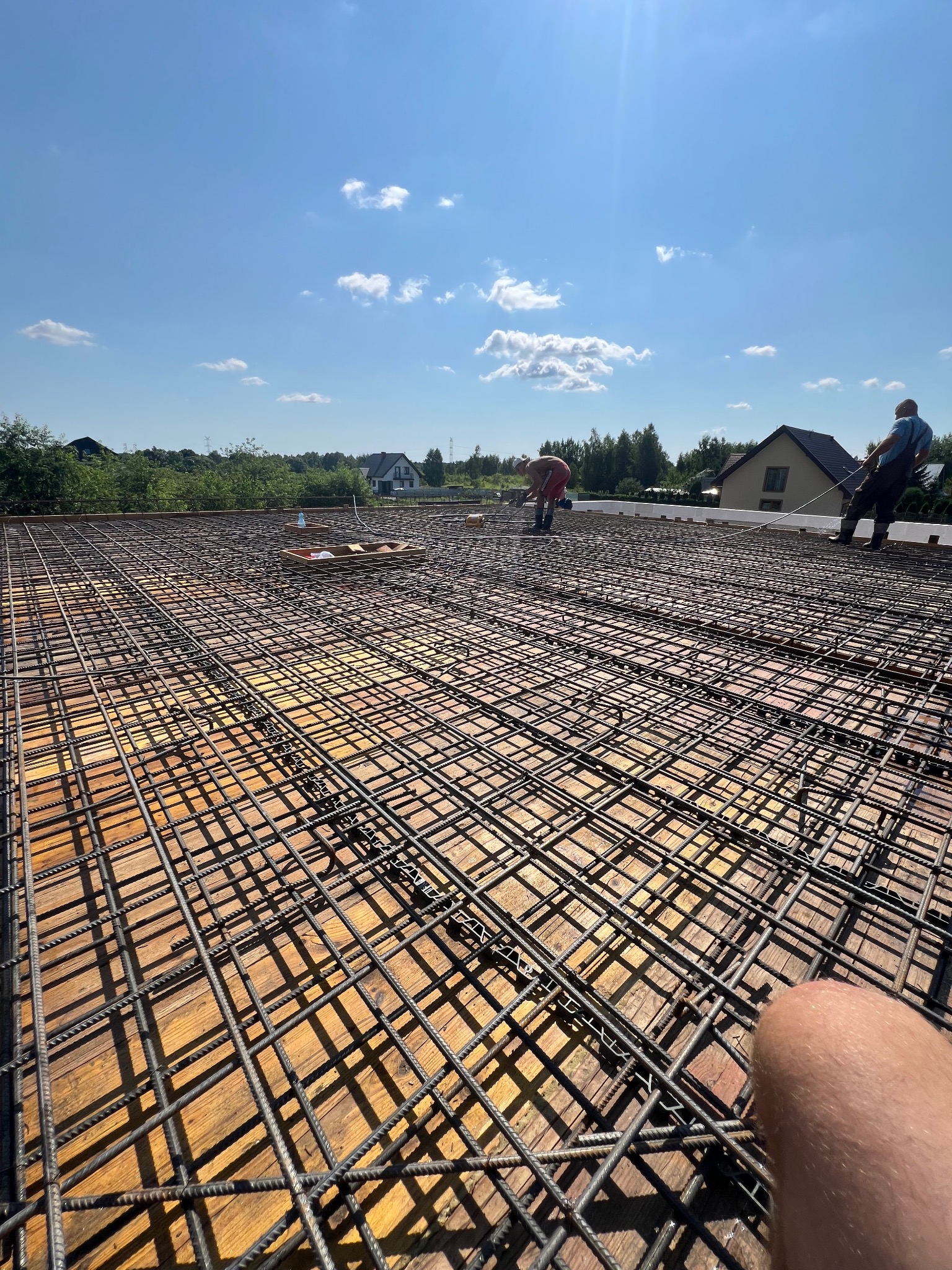 Zbrojenie płyty fundamentowej na drewnianym szalunku, dwóch robotników w oddali przy pracy, widok z góry z fragmentem ciała w kadrze, słoneczny dzień.