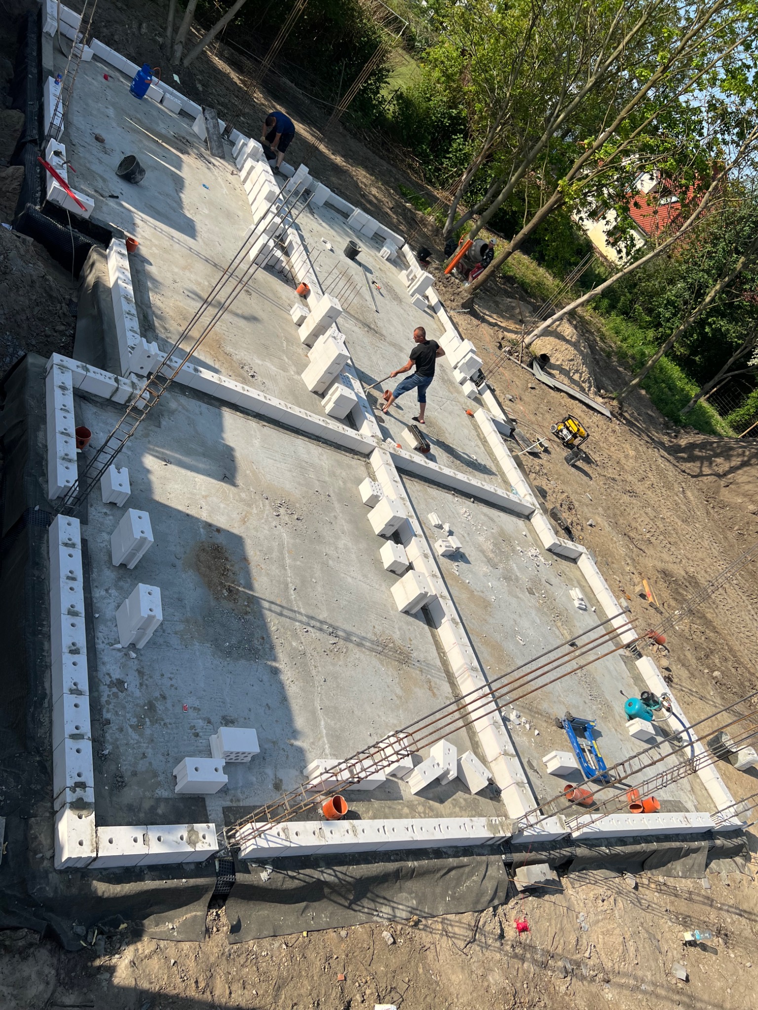 Widok z góry na fundament budynku w trakcie budowy, z pracownikiem układającym bloczki betonowe, widoczne zbrojenia i narzędzia budowlane.