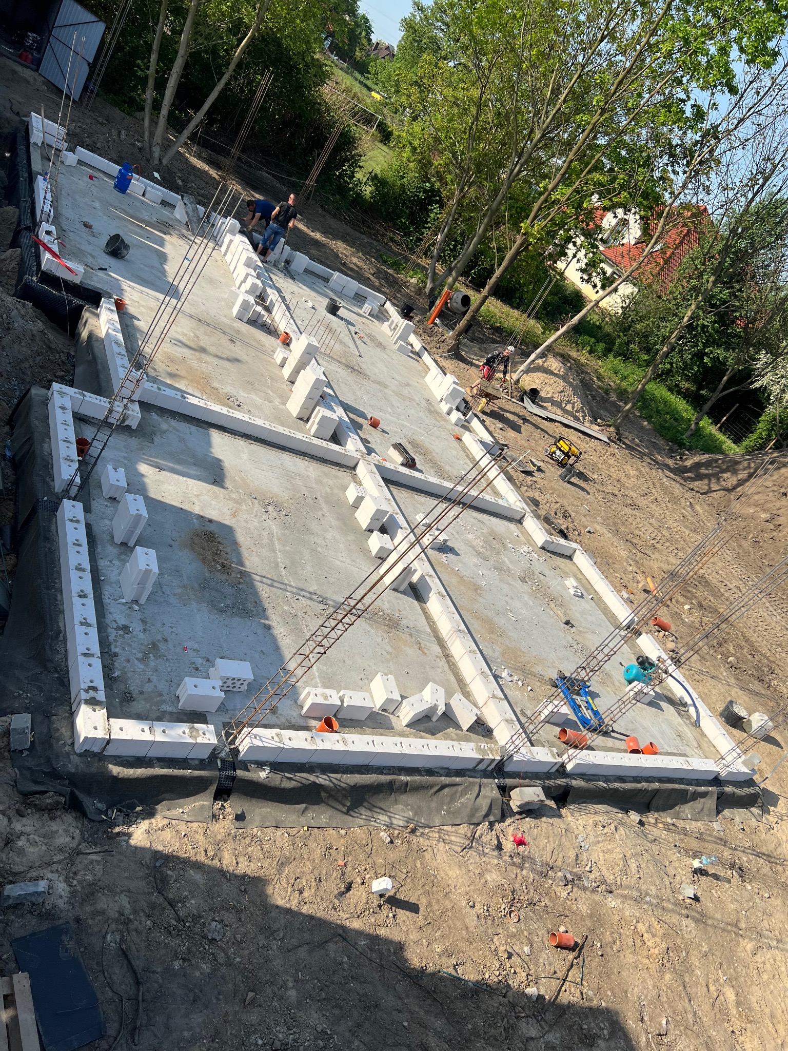 Budowa fundamentów domu: betonowa płyta fundamentowa z ułożonymi bloczkami betonowymi i wystającymi prętami zbrojeniowymi, widoczne narzędzia i pracownicy w tle.