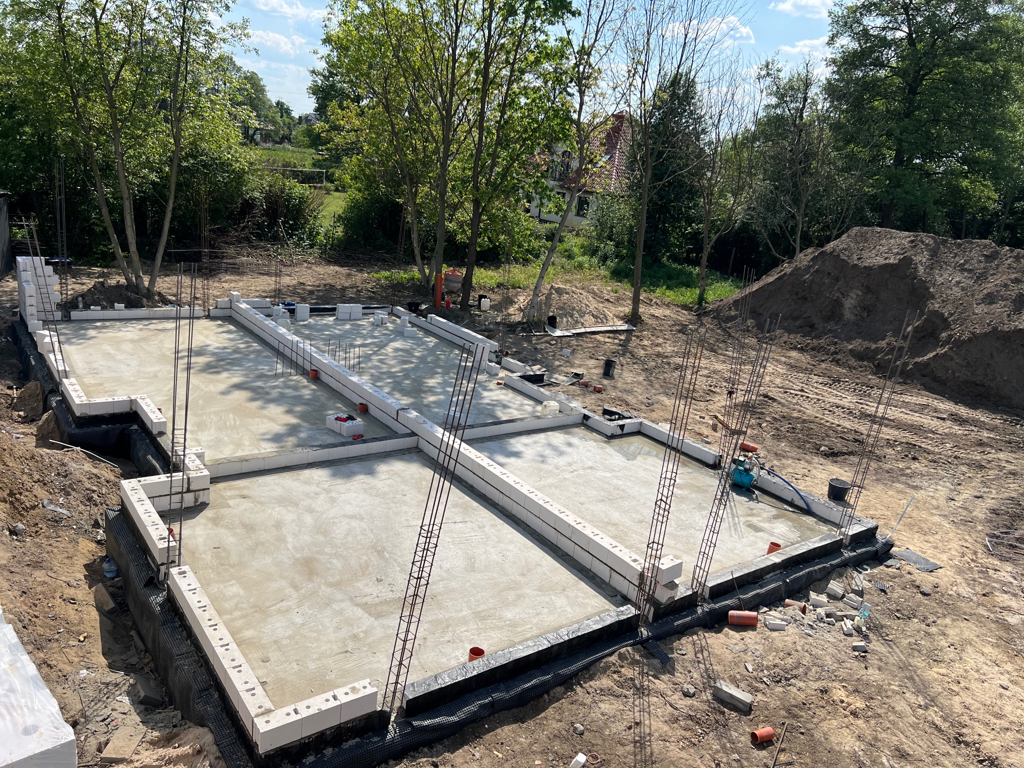 Wylany fundament z betonu z wystającymi prętami zbrojeniowymi, obłożony bloczkami betonowymi, widok z góry na tle drzew i hałdy ziemi.