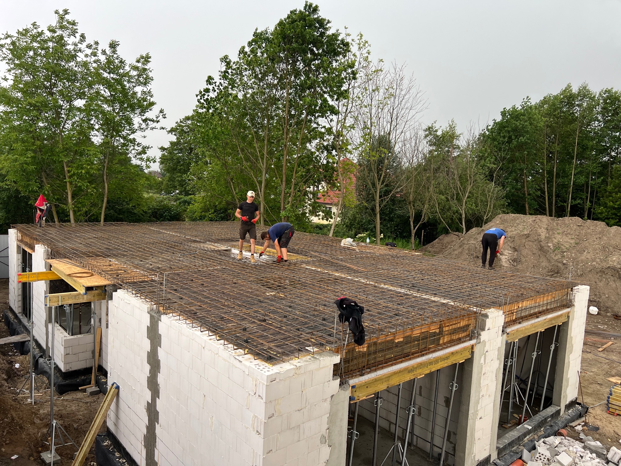 Wiązanie zbrojenia na stropie budynku z białych bloczków, widoczni pracownicy w trakcie pracy, w tle drzewa i hałda ziemi.