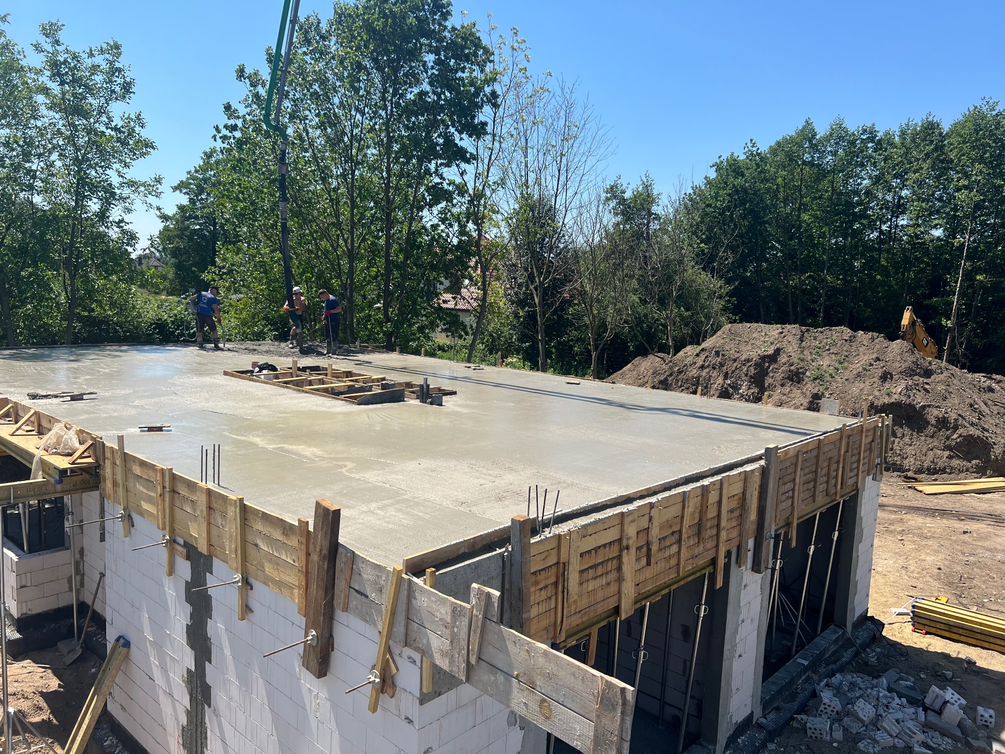 Wylewanie betonu na strop domu z białych bloczków, widoczna pompa do betonu i pracownicy w tle, szalunek z desek na krawędzi stropu.