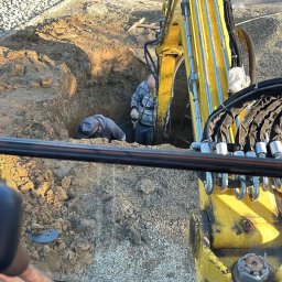 RoxKop - Prace Ziemne Sebastian Górecki - Żółta koparka przy wykopie, dwóch mężczyzn wewnątrz, instalacja paneli słonecznych w tle, widok z kabiny operatora.