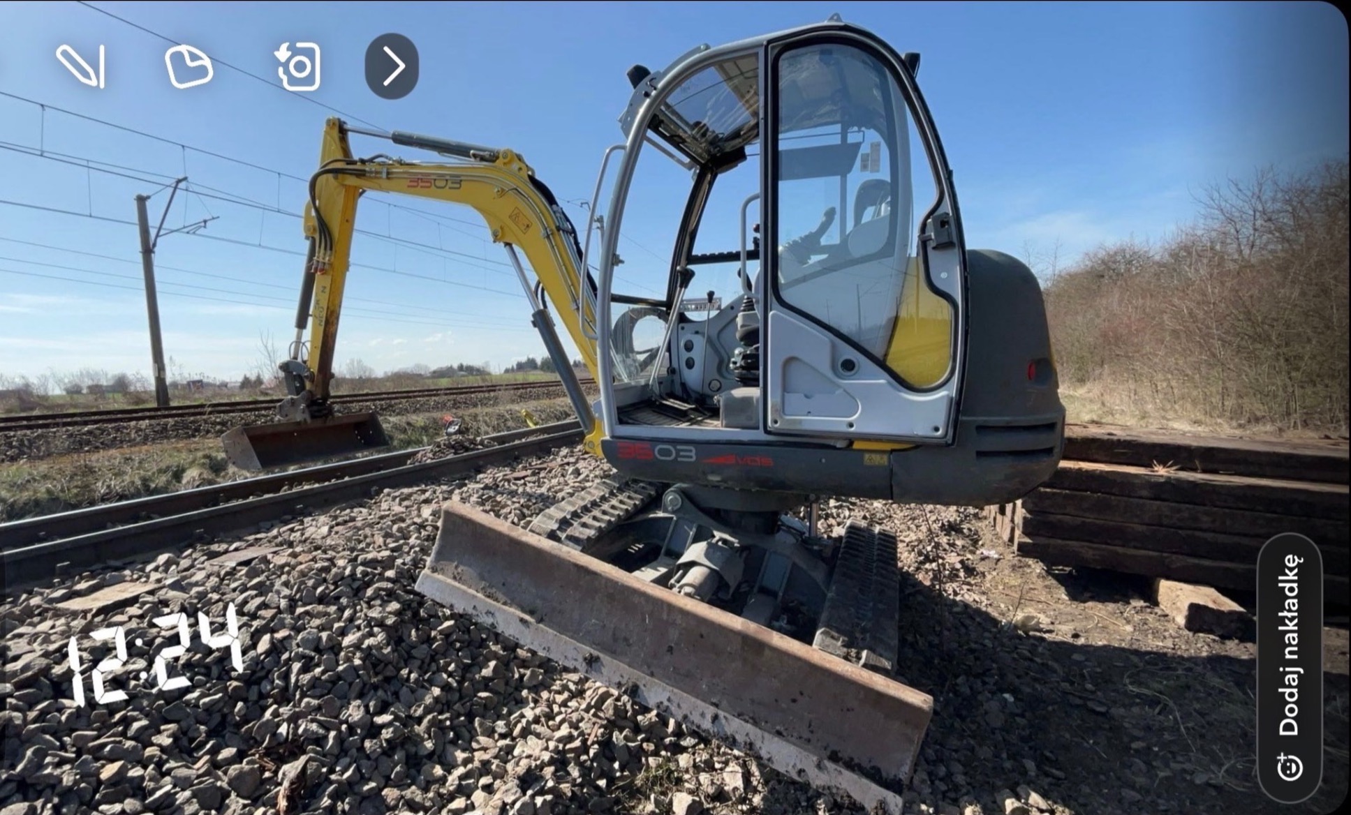 Żółto-szara mini koparka na torach kolejowych, z widocznymi liniami wysokiego napięcia w tle.