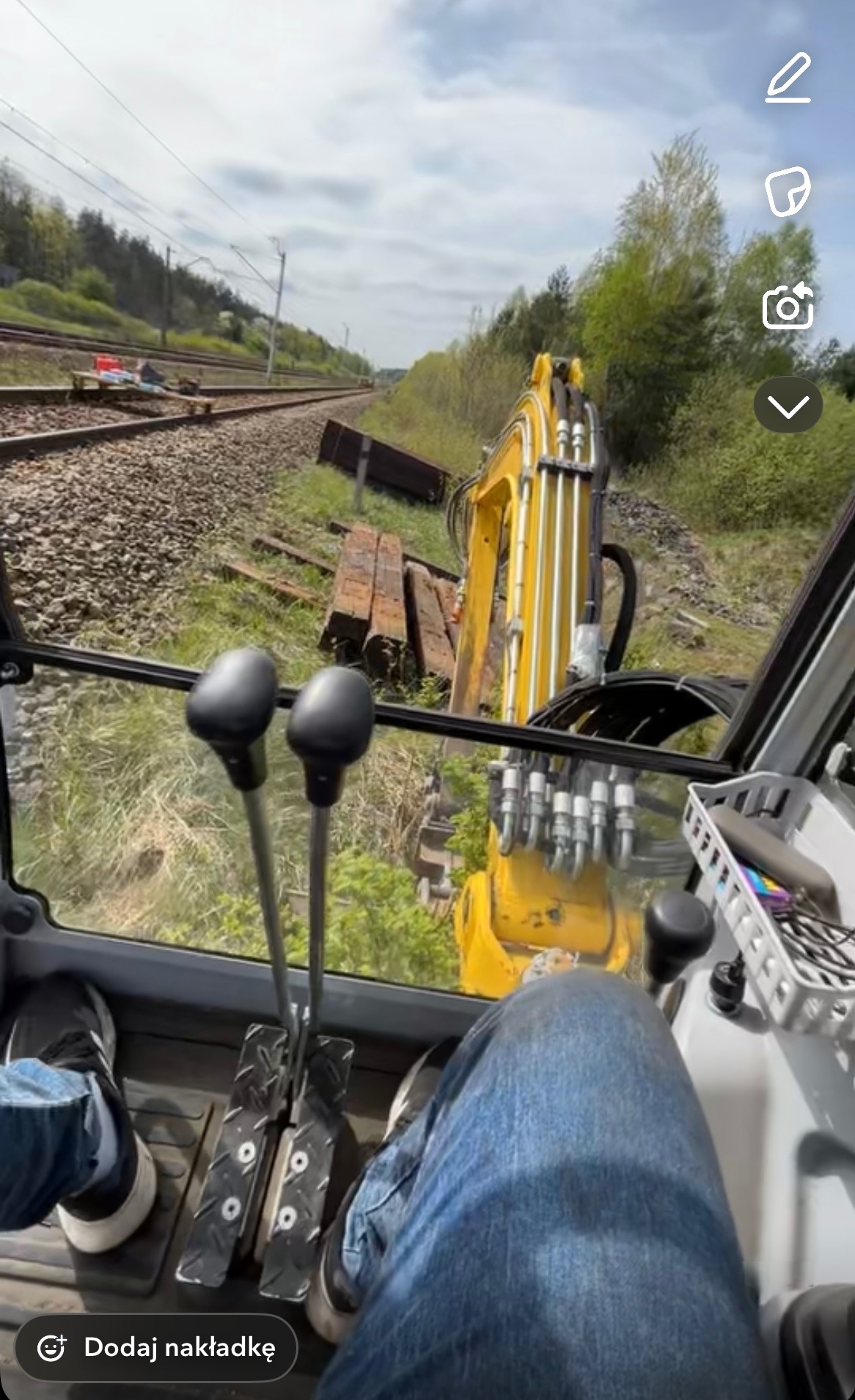 Widok z kabiny operatora żółtej koparki, z widocznymi dżojstikami i pedałami, skierowany na tory kolejowe i brzeg wykopu z drewnianymi podkładami kolejowymi.