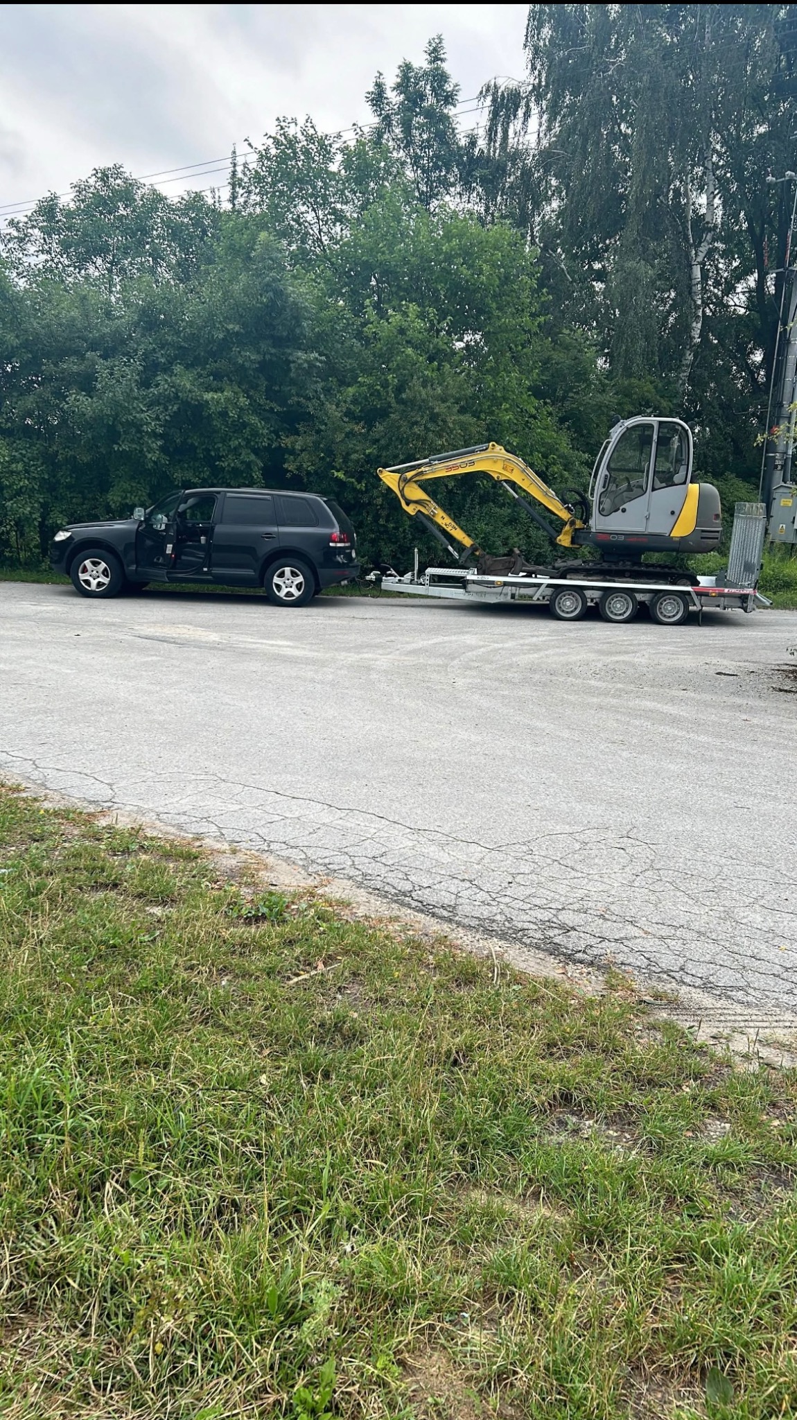 Czarny SUV holuje na przyczepie żółto-szarą mini koparkę na tle zieleni.