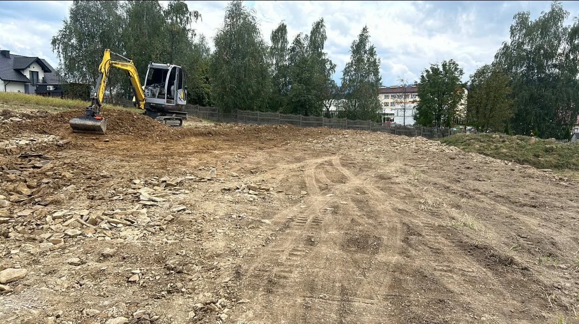 Żółta minikoparka ugniata ziemię na placu budowy; widoczne ślady opon, kamienie i budynki w tle.