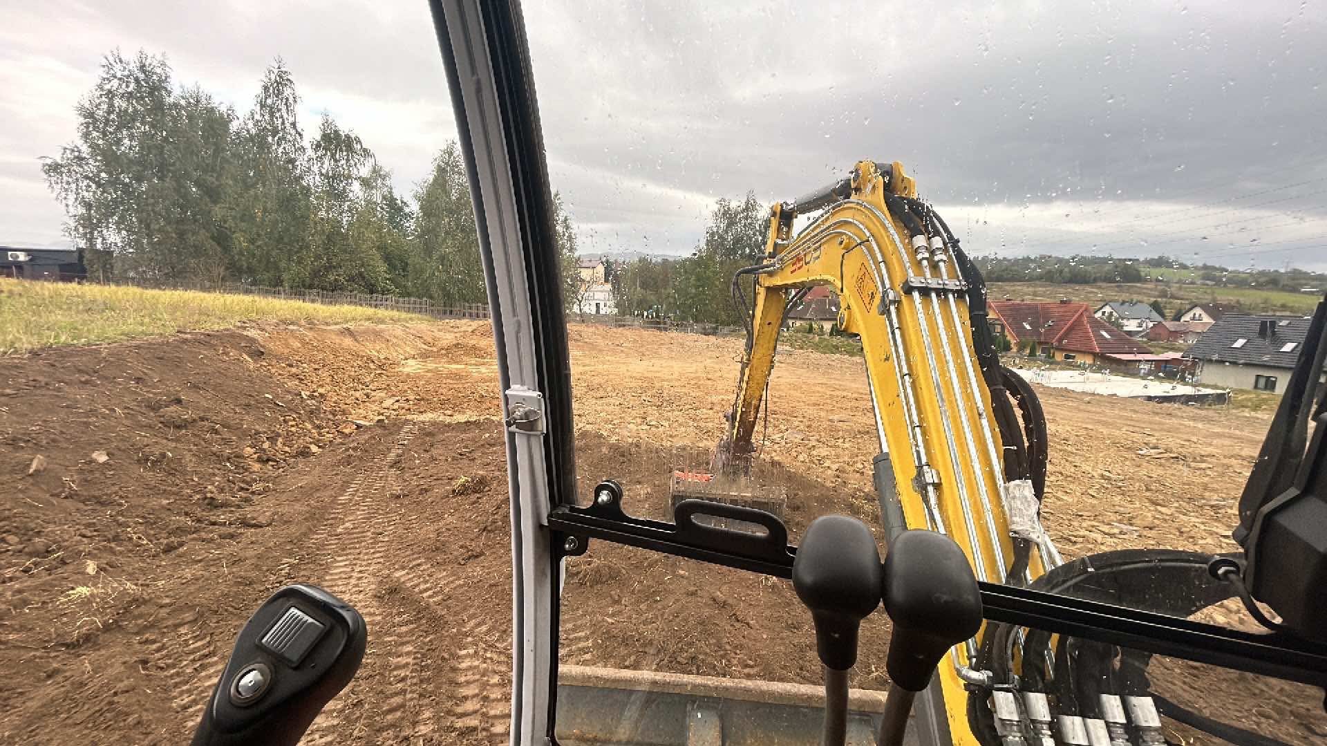 Widok z kabiny operatora na żółtą koparkę pracującą na placu budowy w pochmurny dzień, widoczne ślady opon i domy w tle.