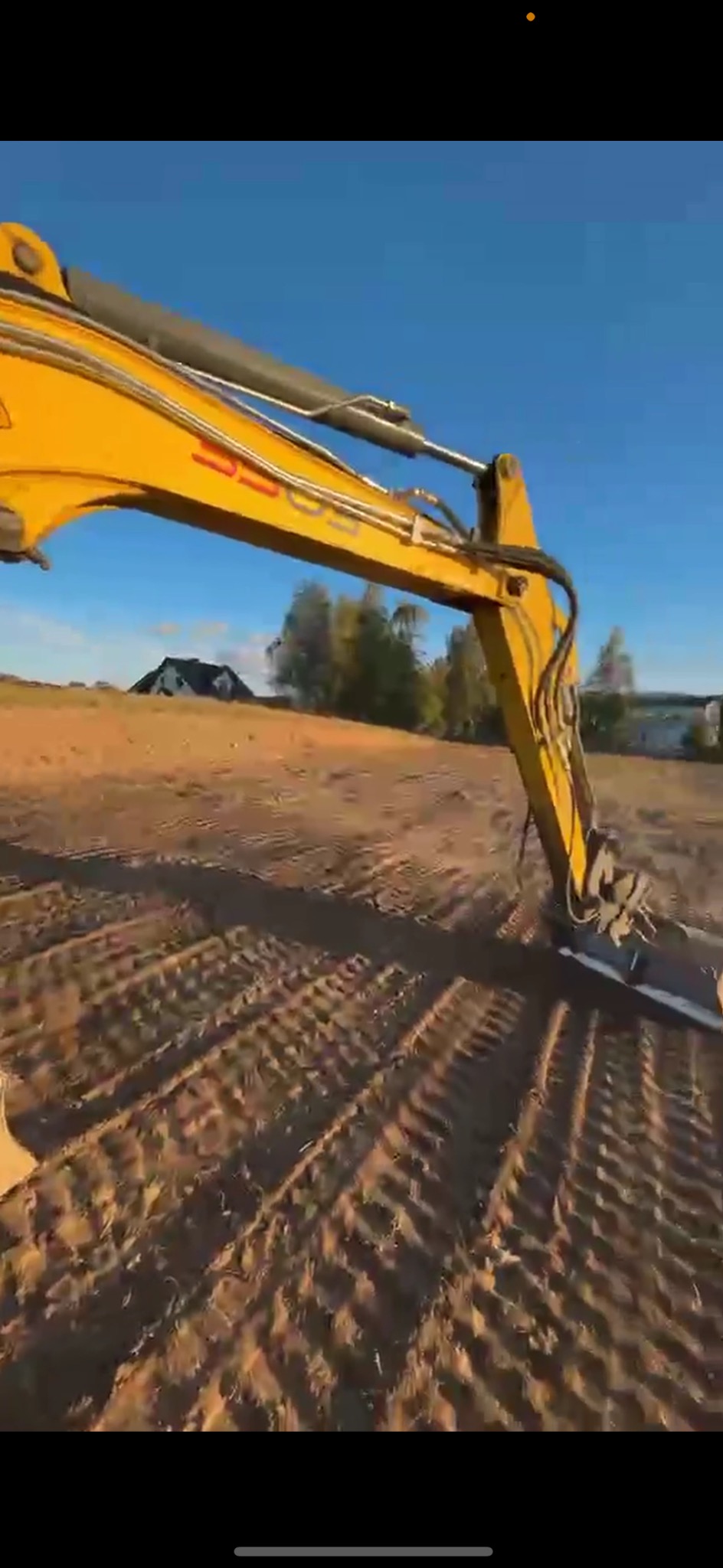 Żółta koparka podczas pracy na polu, widoczne ślady opon na ziemi, w tle domy i drzewa pod błękitnym niebem.