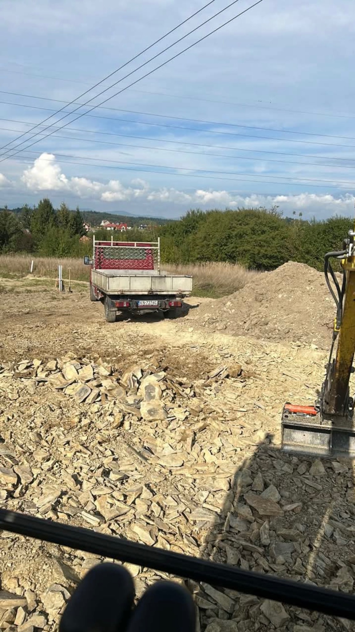 Teren budowy z rozrzuconymi kamieniami, w tle widoczny wywrotka i koparka, krajobraz wiejski z liniami energetycznymi na niebie.