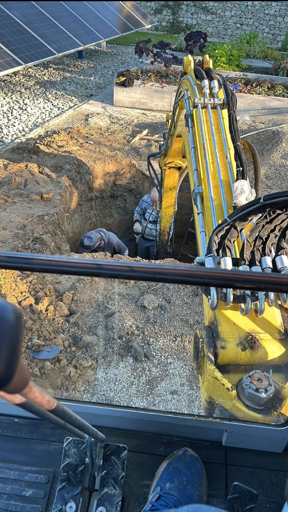 Żółta koparka przy wykopie, dwóch mężczyzn wewnątrz, instalacja paneli słonecznych w tle, widok z kabiny operatora.