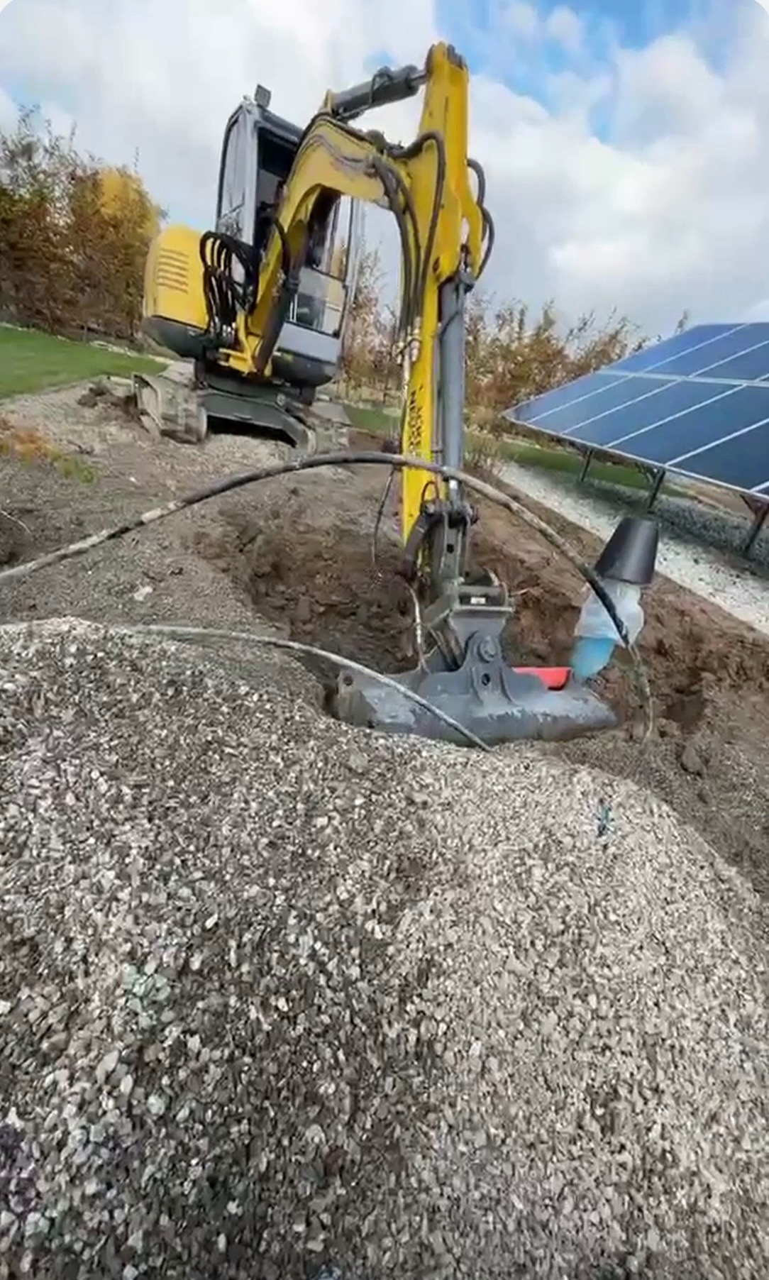 Żółta koparka w trakcie pracy, zagłębiająca łyżkę w ziemię, obok widoczne panele słoneczne i hałda żwiru.