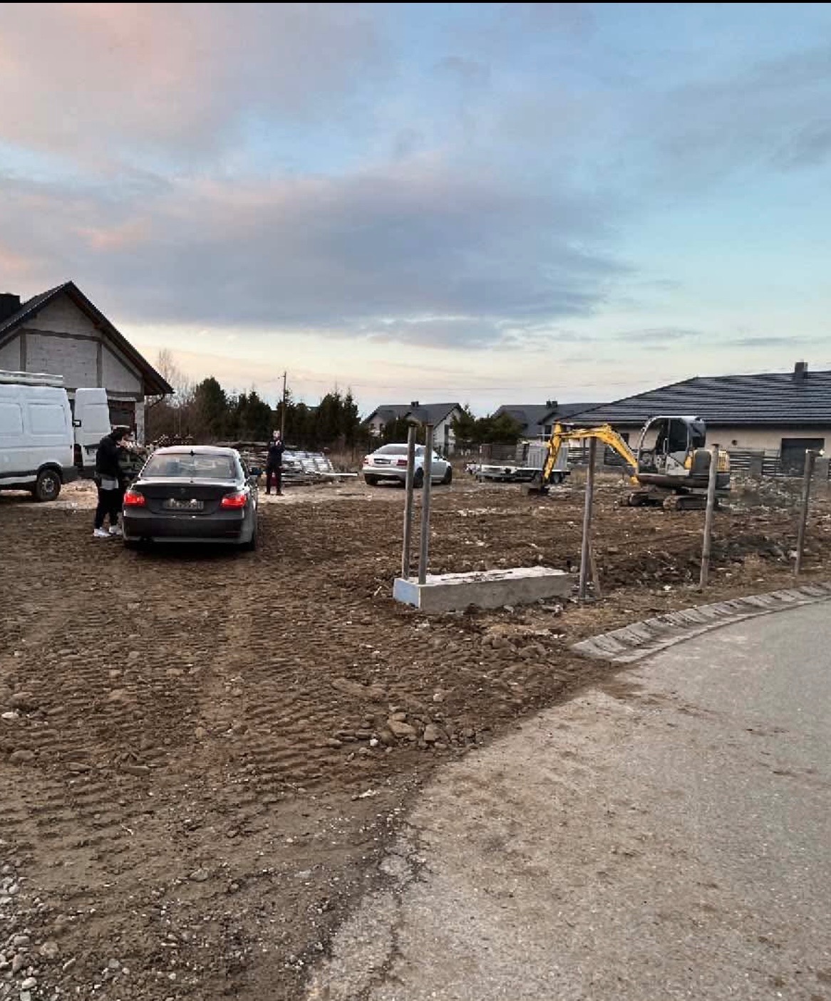 Żółta mini koparka na placu budowy z widocznymi fundamentami, samochodem osobowym i dostawczym w tle, pod pochmurnym niebem.