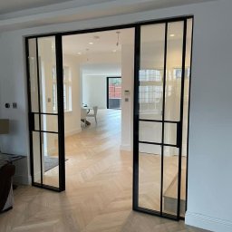 Metalovo - Nowoczesne, przesuwne drzwi wewnętrzne z czarnymi szprosami i przezroczystym szkłem, na tle parkietu ułożonego w jodełkę. Widok na jasne wnętrze domu.