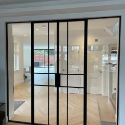 Metalovo - Nowoczesne, dwuskrzydłowe drzwi wewnętrzne z czarnymi szprosami i przezroczystym szkłem, oddzielające pomieszczenia z parkietem ułożonym w jodełkę.