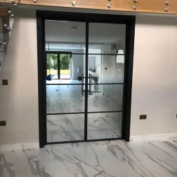 Metalovo - Nowoczesne, dwuskrzydłowe drzwi szklane z czarną ramą, widoczne na tle marmurowej podłogi i jasnych ścian. Minimalistyczny design z widocznymi elementami mocującymi.