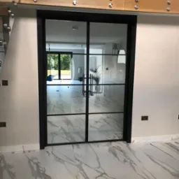 Nowoczesne, dwuskrzydłowe drzwi szklane z czarną ramą, widoczne na tle marmurowej podłogi i jasnych ścian. Minimalistyczny design z widocznymi elementami mocującymi.